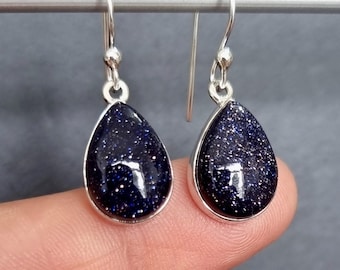 May include: A pair of silver-toned drop earrings. Each earring features a teardrop-shaped, dark blue stone with sparkling inclusions, set in a silver bezel. The earrings hang from a simple hook.