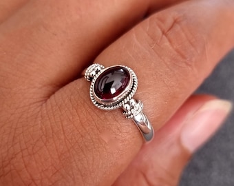May include: A silver ring with a red oval gemstone set in a decorative bezel. The ring has a delicate band and a detailed design.