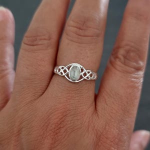 May include: A silver ring with a light-colored oval gemstone. The ring features a Celtic knot design on the band, with the gemstone set in the center. The ring is on a finger, with a dark background.