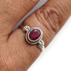 May include: A silver ring featuring a red oval gemstone set in an ornate bezel. The ring has a textured band and small decorative accents on either side of the stone.
