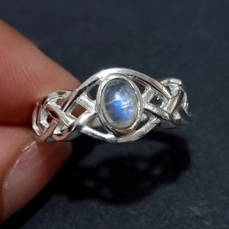 May include: A silver ring with a Celtic knot design. The ring features an oval-shaped, iridescent moonstone in the center. The band has an intricate woven pattern. The ring is displayed against a dark background.