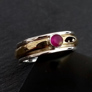 Peut inclure: Une bague en argent et or avec une pierre précieuse rouge sertie au centre. La bague a une bande texturée.