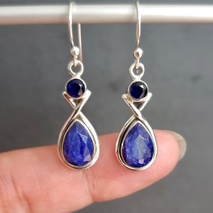 May include: A pair of silver dangle earrings with blue gemstone drops. The earrings feature a teardrop-shaped gemstone with a smaller round gemstone at the top.