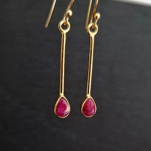 Könnte beinhalten: Ein Paar goldfarbene Ohrhänger mit tropfenförmigen, tiefroten Edelsteinen. Die Ohrringe haben ein schlichtes, elegantes Design mit einem langen, schlanken Goldstab und einem kleinen, runden Akzent oben.
