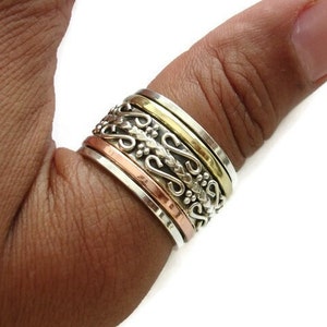 May include: A silver, copper, and brass spinner ring with an intricate design. The ring is worn on a person's finger.