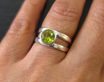 May include: A silver ring featuring a round, faceted, lime-green peridot gemstone set in a bezel. The band is split into two parallel silver bars that connect at the base, creating an open space between the two bands on either side of the stone.