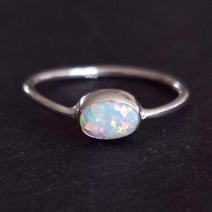 Può includere: Un anello in argento con una fascia liscia e lucida e una pietra di opale di forma ovale al centro. L'opale mostra bagliori iridescenti di luce rosa, verde e blu su una base bianco latte, incastonato in una semplice montatura.