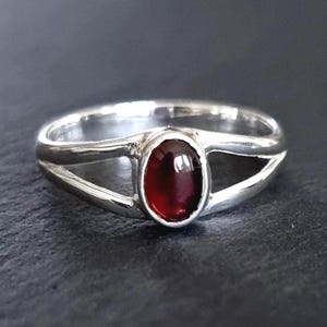 May include: A silver ring featuring a polished, oval-shaped deep red garnet gemstone set in the centre. The band is split into two separate silver strands that meet at the stone setting, creating an open, bypass-style design with a smooth finish.