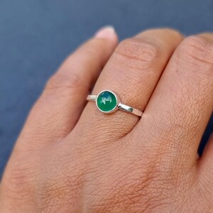 May include: A silver ring featuring a small, round, polished green gemstone set in a simple metal bezel. The band is thin and smooth, showcasing a minimalist design that highlights the vibrant green color of the stone against the metallic silver.
