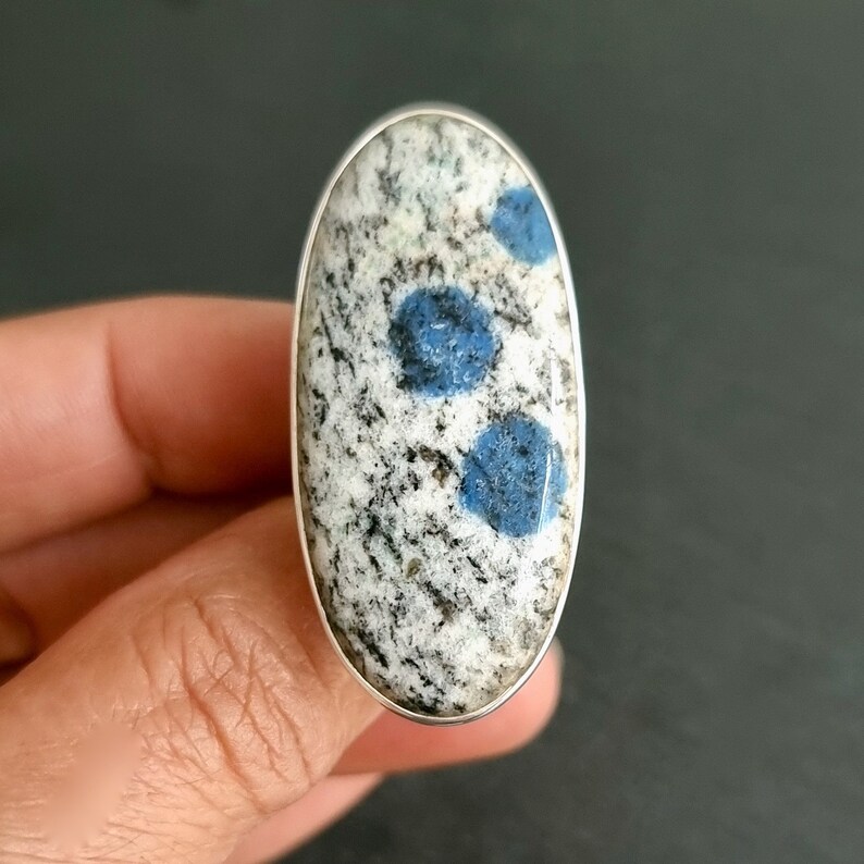 May include: An oval-shaped ring featuring a polished stone with a speckled white, gray, and black granite-like texture, accented by three distinct, circular, vibrant blue inclusions set within a smooth, thin silver metal band frame.
