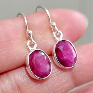 Peut inclure: Une paire de boucles d'oreilles pendantes avec des pierres précieuses rubis rose foncé de forme ovale et facettées, serties dans des chatons en argent poli. Chaque boucle est suspendue à un crochet en argent simple et élégant.