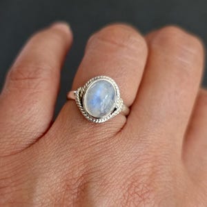 May include: A silver ring featuring an oval-shaped moonstone cabochon with a blue iridescent sheen. The stone is set in a polished silver bezel, surrounded by a decorative twisted rope-style metal band that encircles the central gemstone setting.