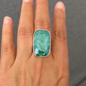 Può includere: Un anello rettangolare con taglio smeraldo con una grande gemma verde vibrante incastonata in una fascia d'argento. La gemma presenta inclusioni e sfaccettature visibili, che riflettono la luce. L'anello è indossato su un dito.