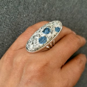 May include: An oval-shaped ring featuring a polished K2 granite stone set in a silver-toned metal band. The stone has a speckled white and grey base with distinct, circular, vibrant blue inclusions scattered across the smooth, rounded surface.