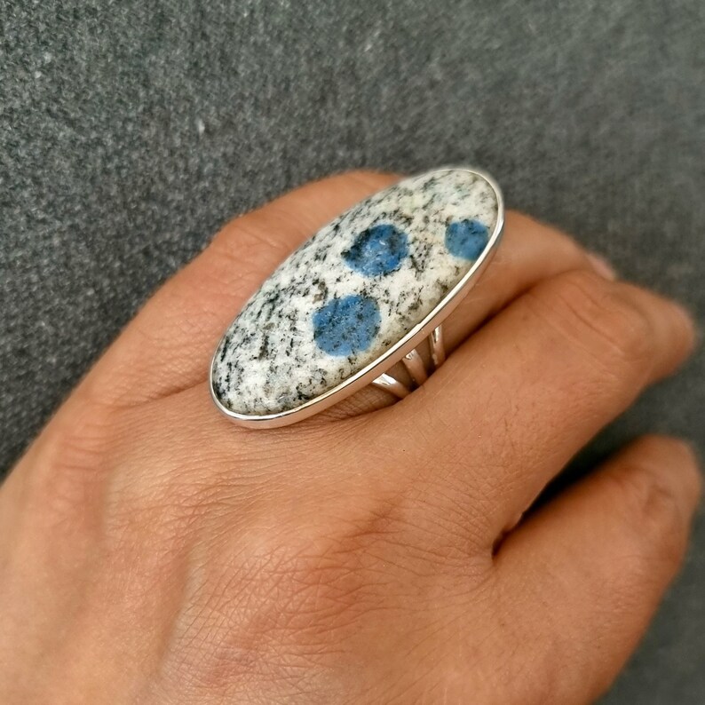 May include: An oval-shaped ring featuring a polished K2 granite stone set in a silver-toned metal band. The stone has a speckled white and grey base with distinct, circular, vibrant blue inclusions scattered across the smooth, rounded surface.