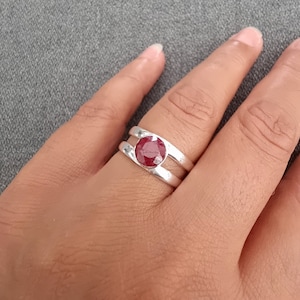 Könnte beinhalten: Ein silberner Ring mit einem roten Edelstein in der Mitte. Der Ring hat ein Doppelband-Design.