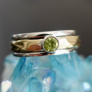 May include: A silver and gold ring featuring a green gemstone. The ring is crafted from three bands, with the top band being gold and the bottom two bands being silver.
