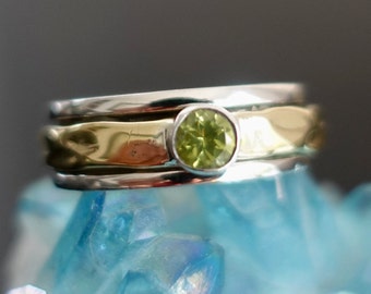 Könnte beinhalten: Ein Silber- und Goldring mit einem grünen Edelstein. Der Ring besteht aus drei Bändern, wobei das obere Band aus Gold und die beiden unteren Bänder aus Silber sind.