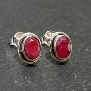 May include: A pair of silver earrings featuring red ruby gemstones. The earrings are oval shaped and have a smooth, polished finish.
