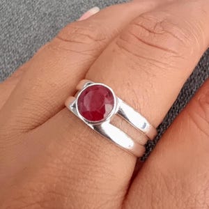 Peut inclure: Une bague en argent ornée d'une pierre précieuse rouge foncé, ronde et à facettes, montée dans un serti clos. L'anneau présente une conception fendue, où deux tiges d'argent distinctes convergent à la base, créant un espace ouvert.