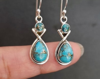 Può includere: Un paio di orecchini pendenti in argento con una pietra turchese a forma di goccia con venature color rame alla base e una pietra turchese rotonda più piccola sopra, collegate da una montatura in argento intrecciato e un gancio.