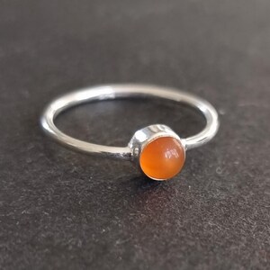 May include: A silver ring featuring a round, orange gemstone. The gemstone is set in a simple bezel setting, with a thin, polished band. The ring is displayed against a dark backdrop.