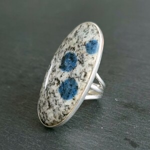 May include: An oval-shaped ring featuring a polished K2 stone set in a silver band. The stone has a speckled white and grey granite-like appearance with three distinct, circular, vibrant blue spots embedded across its smooth surface.