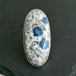 May include: An oval-shaped ring featuring a polished stone cabochon set in a smooth silver bezel. The stone has a speckled white and grey granite-like base with three distinct, circular, vibrant blue inclusions embedded across the surface.