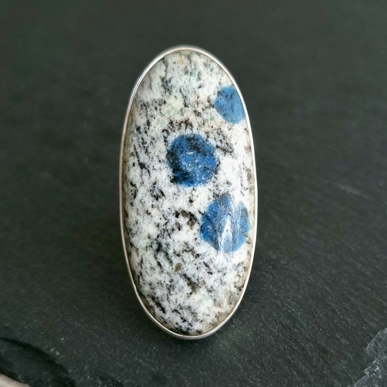 May include: An oval-shaped ring featuring a polished stone cabochon set in a smooth silver bezel. The stone has a speckled white and grey granite-like base with three distinct, circular, vibrant blue inclusions embedded across the surface.