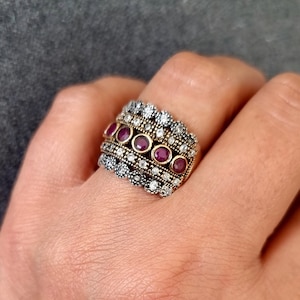 May include: A silver and gold ring with a band of small white stones and a row of six red stones.