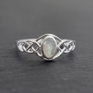 May include: A silver ring with an oval, iridescent gemstone set in a Celtic knot design. The ring's band features intricate knotwork on either side of the gemstone, creating a unique and elegant piece of jewelry.