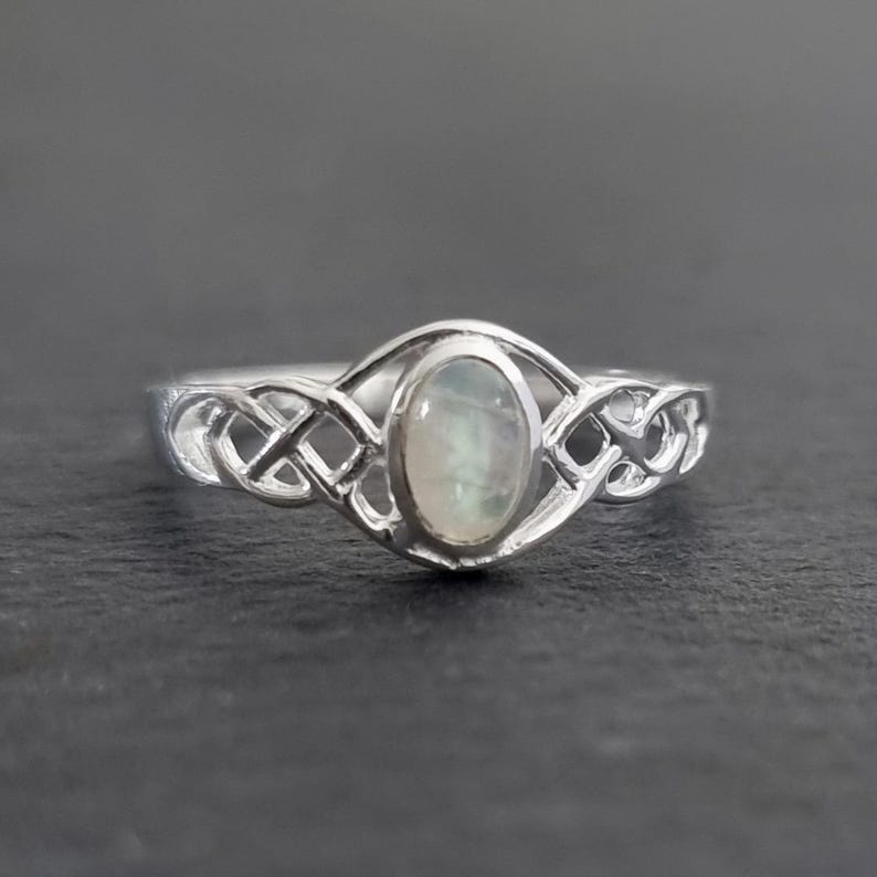 May include: A silver ring with an oval, iridescent gemstone set in a Celtic knot design. The ring's band features intricate knotwork on either side of the gemstone, creating a unique and elegant piece of jewelry.