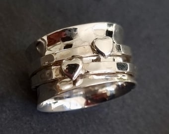 Könnte beinhalten: Ein drehbarer Silberring mit drei Bändern und zwei herzförmigen Akzenten. Der Ring ist so konzipiert, dass er am Finger frei drehbar ist.