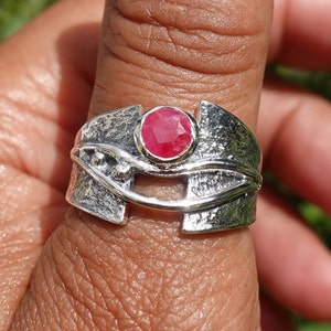 May include: A silver ring with a textured design features a bright red gemstone set in the centre. The ring is worn on a finger.