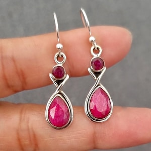 May include: Silver drop earrings with ruby gemstones. Each earring has a small round gem, a decorative silver element, and a larger teardrop-shaped gem. The earrings are shown against a neutral background. The gemstones are a deep red colour.