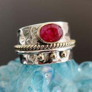 May include: A silver ring with a red gemstone set in a gold bezel. The ring has a textured band and a gold rope design around the top.