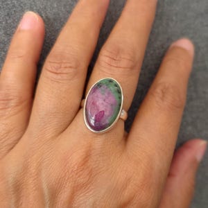 May include: Oval-shaped ring featuring a large, polished gemstone with a mix of pink, green, and white colors. The gemstone is set in a silver band. The ring is displayed on a hand against a gray background.