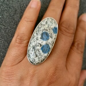 May include: An oval-shaped ring featuring a polished stone with a speckled white and black granite-like texture, accented by two distinct circular blue inclusions. The stone is set within a smooth, thin silver-toned metal bezel frame.