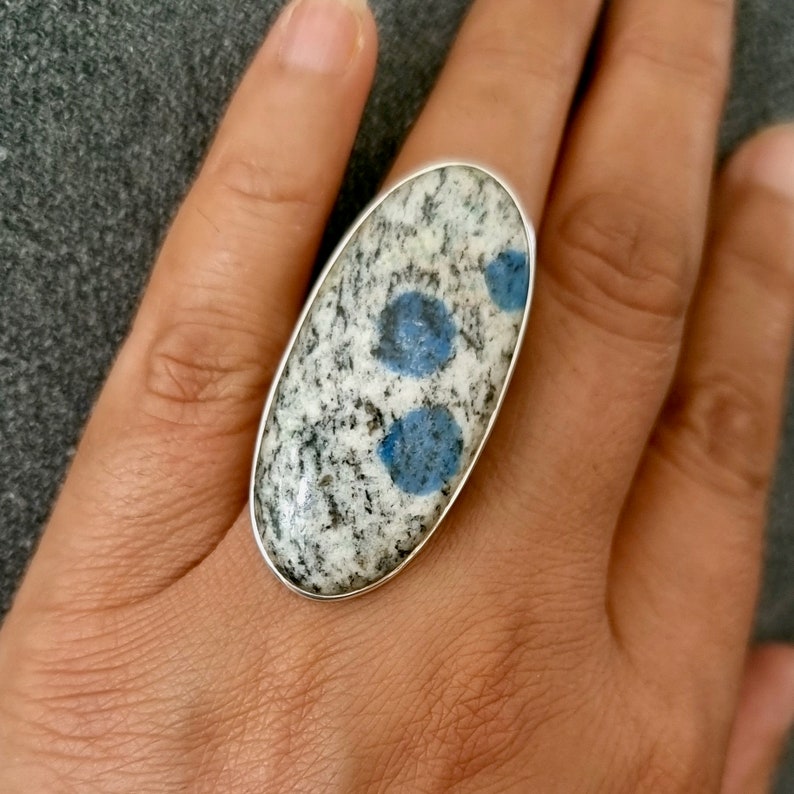 May include: An oval-shaped ring featuring a polished stone with a speckled white and black granite-like texture, accented by two distinct circular blue inclusions. The stone is set within a smooth, thin silver-toned metal bezel frame.