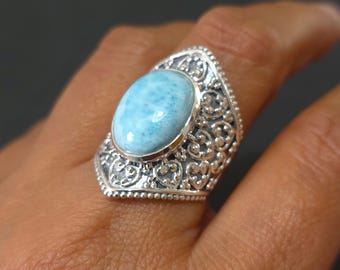May include: A silver ring with an oval, light blue gemstone. The ring has an ornate, filigree design around the stone and along the band. The gemstone is approximately 2 cm long.