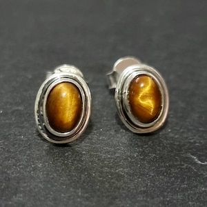 Pode incluir: Um par de brincos de prata com pedras de olho de tigre de forma oval. Os brincos têm uma configuração de bezel de prata e um fecho de poste.