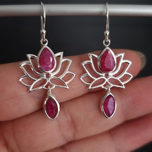 Könnte beinhalten: Ein Paar silberne Ohrringe mit roten Edelstein-Lotusblüten-Designs. Die Ohrringe haben einen baumelnden Tropfen mit einem roten Edelstein.