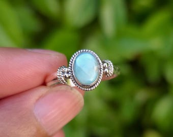 May include: A silver ring featuring an oval, light blue gemstone. The gemstone is surrounded by a twisted rope design and decorative accents on the sides. The ring is held up against a blurred green background.