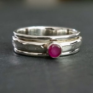 May include: A silver ring with a pink gemstone set in the centre. The ring has a textured band and is made of three stacked bands.