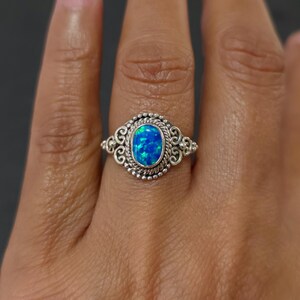 May include: A silver ring with an oval, blue opal stone. The opal is surrounded by a detailed, ornate silver setting with a beaded border and scrollwork accents. The ring is shown on a person's finger.