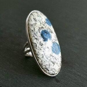 May include: A silver ring featuring a large, oval-shaped stone with a speckled white and gray granite-like texture, accented by three distinct, rounded blue inclusions set within a smooth, polished silver bezel and a split-shank band.