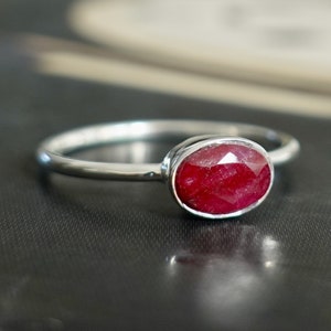 May include: A silver ring with a vivid, oval-cut ruby gemstone. The ruby is a deep red hue, set in a bezel. The band is slender and polished, offering a classic and elegant appearance. The ring is suitable for everyday wear.