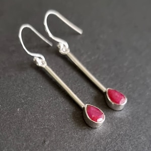May include: A pair of silver drop earrings with a teardrop-shaped, deep red gemstone at the bottom of each earring. The earrings have a simple, elegant design with a hook closure.