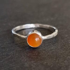 Può includere: Anello d'argento con una gemma arancione. L'anello ha una fascia martellata e un design semplice. La gemma è rotonda e lucida, incastonata in uno stile minimalista. Il design dell'anello è elegante e sobrio.