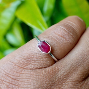 May include: A silver ring with a vivid oval-cut ruby gemstone. The ruby is a deep red hue, set in a simple, elegant band. The ring is displayed against a blurred green backdrop.